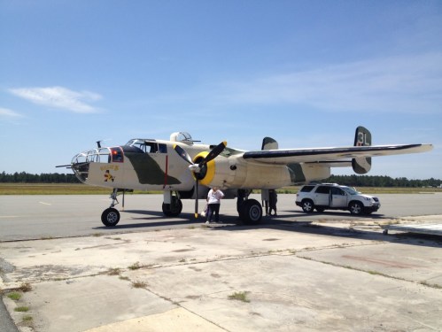 A vintage military aircraft parked on an airstrip, with a person standing nearby and a vehicle in the background.