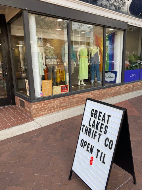 A thrift store window display featuring mannequins in colorful clothing, with a sign reading "Great Lakes Thrift Co. Open til 8."