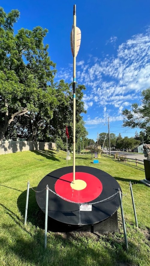 A large dartboard with a red bullseye and an arrow stuck in the center, set on a grassy area under a blue sky.
