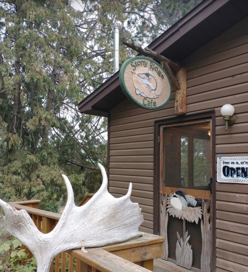 Sign for Stony Ridge Cafe on a wooden building, featuring a fish design and a large moose antler sculpture nearby.