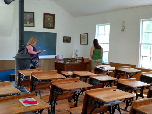 Two women explore a historic classroom with wooden desks, a chalkboard, and vintage decor.