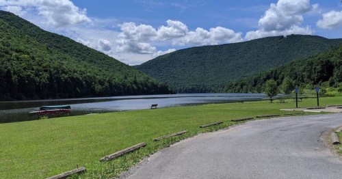 A serene lake surrounded by green mountains under a partly cloudy sky, with a grassy area and a winding road.