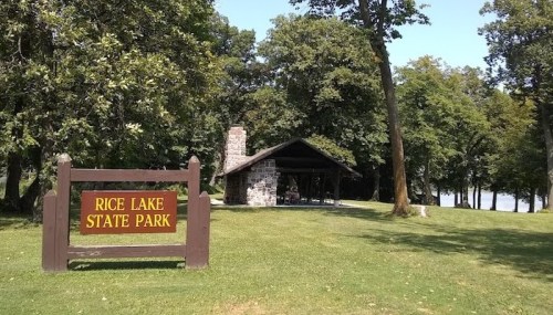A stone shelter at Rice Lake State Park, surrounded by trees and grass, with a sign in front.