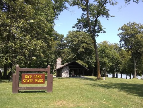 A sign for Rice Lake State Park in front of a stone shelter surrounded by trees and a lake in the background.