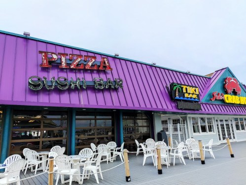 Colorful restaurant exterior featuring signs for a pizza place, sushi bar, tiki bar, and crab house with empty chairs outside.