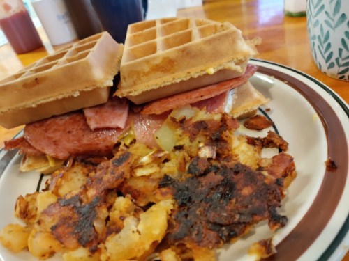 A plate with a waffle sandwich filled with meat and cheese, alongside crispy, browned hash browns.