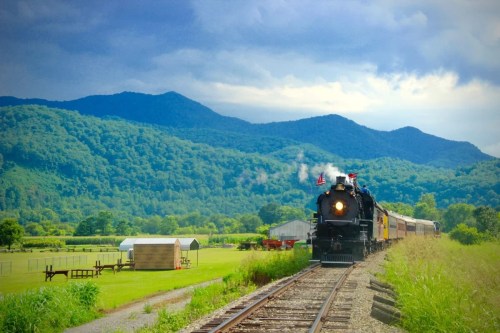 A vintage steam train travels along tracks with mountains and a farm in the background under a cloudy sky.