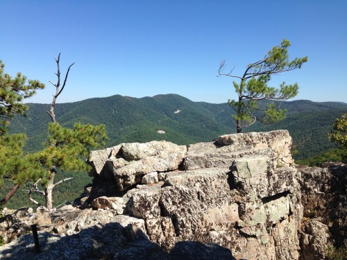 A rocky outcrop overlooks a lush green valley and distant mountains under a clear blue sky.