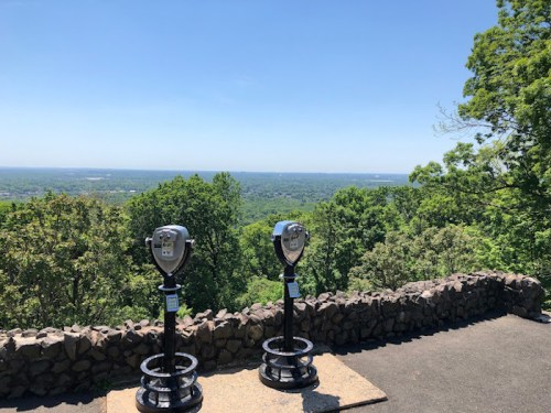 Two viewing binoculars on a stone platform overlooking a lush green landscape under a clear blue sky.
