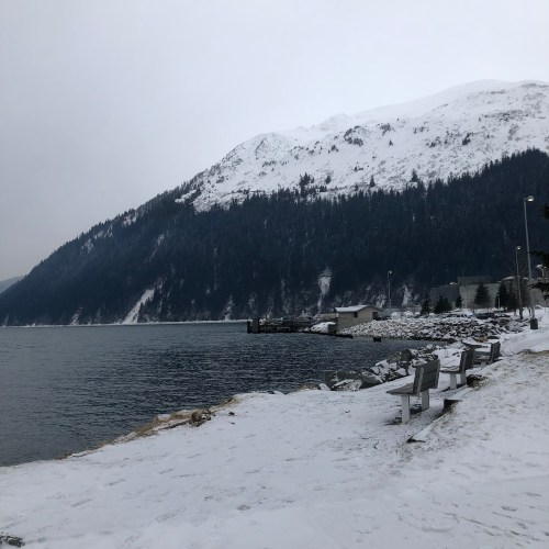 Snow-covered landscape by a calm lake, with mountains in the background and benches along the shore.