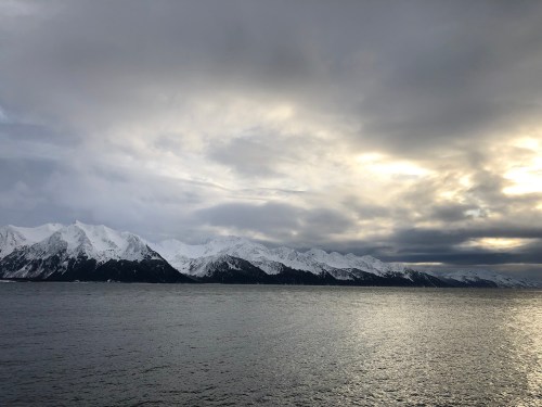 A serene view of snow-capped mountains under a cloudy sky, reflecting on calm waters.