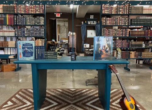 A cozy bookstore with a blue table, bookshelves filled with books, and a guitar on the floor. Warm, inviting atmosphere.