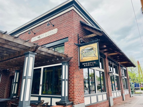 Exterior of a brick brewpub and grill with a sign, featuring a covered patio and cloudy sky.