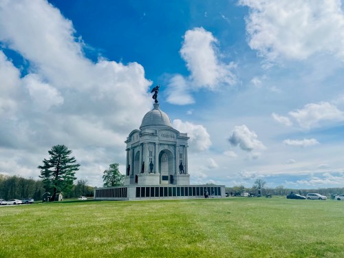 A large white monument with a dome and statue on top, surrounded by green grass and a cloudy blue sky.