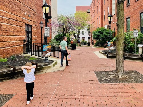 A child walks ahead on a brick path while an adult follows, surrounded by buildings and greenery.