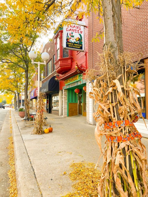 A colorful street scene with autumn leaves, featuring a restaurant and decorated corn stalks along the sidewalk.