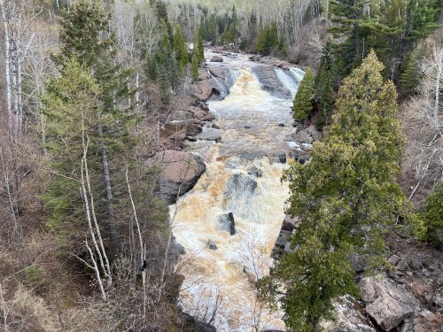 A rushing river cascades over rocky terrain, surrounded by lush green trees and a serene natural landscape.