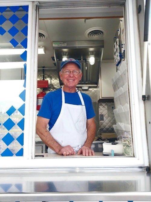 A smiling older man in a blue shirt and white apron stands at a food truck window, ready to serve customers.