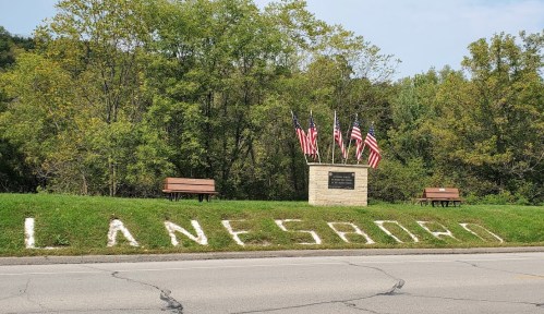 Sign reading "Lanesboro" with benches and American flags in a green, tree-lined area near a road.