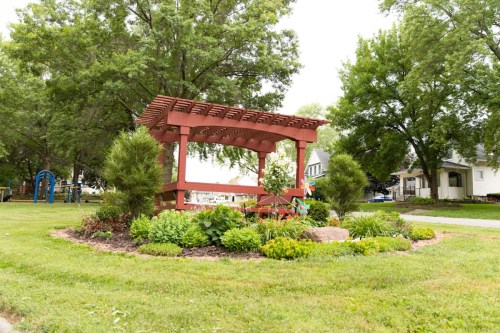 A red wooden pergola surrounded by colorful flowers and greenery in a park setting.