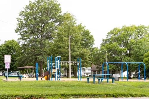A playground with blue equipment, swings, and benches, surrounded by green trees and grass on a cloudy day.