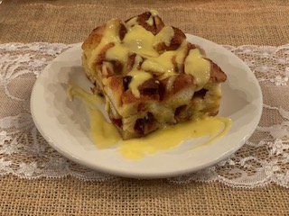 A slice of bread pudding drizzled with creamy sauce on a white plate, set on a textured burlap background.