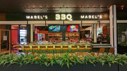 A modern BBQ restaurant interior featuring a bar, seating, and large screens displaying sports. Green plants line the front.