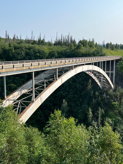 A large arch bridge spans a lush green valley, surrounded by trees under a clear blue sky.