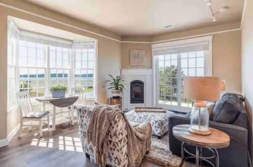 Cozy living room with large windows, a fireplace, and stylish furniture, offering a view of the outdoors.