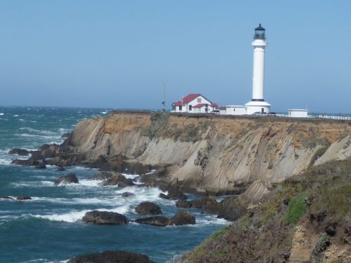 A white lighthouse stands on a rocky cliff by the ocean, with waves crashing against the shore under a clear blue sky.