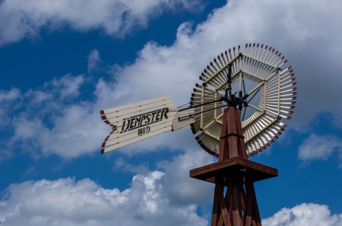 A vintage windmill with a sign reading "Denster 1929" against a backdrop of blue sky and fluffy white clouds.