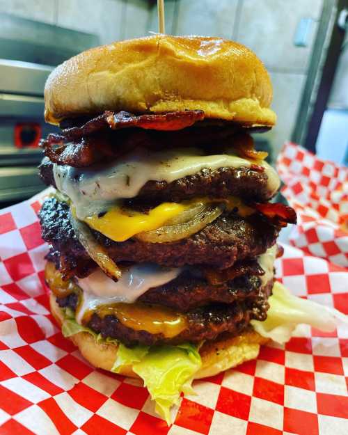 A towering burger stacked with multiple patties, bacon, cheese, and onions, served on a red and white checkered paper.