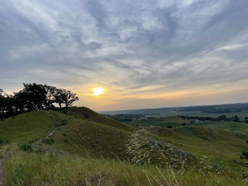 A serene landscape at sunset, featuring rolling hills, a tree line, and a colorful sky with clouds.
