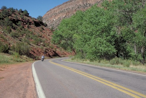 A winding road through a scenic landscape with red rock formations and green trees under a clear blue sky.