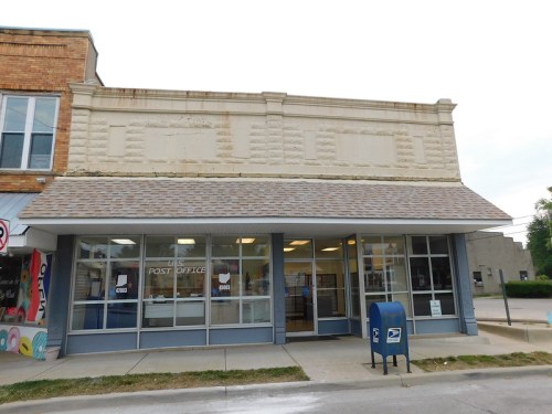 A small post office building with a flat roof and large windows, featuring a blue mailbox in front.