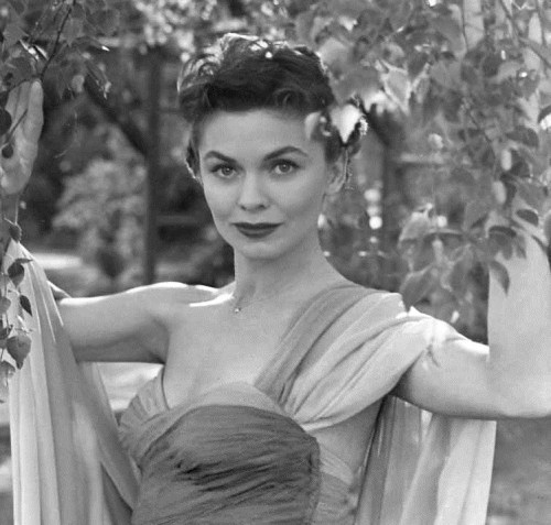 A woman in a flowing dress poses gracefully among greenery, exuding elegance and charm in a black-and-white photo.