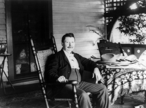 A man with a mustache sits in a rocking chair on a porch, surrounded by books and papers on a table.
