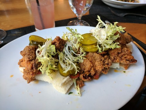 A plate of fried chicken on a bun, topped with pickles, lettuce, and sliced onions, served with a drink in the background.