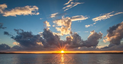 A vibrant sunset over a calm lake, with rays of light shining through clouds and reflecting on the water.
