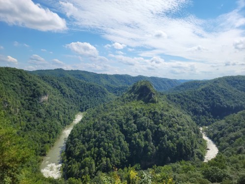 A scenic view of lush green hills and a winding river under a bright blue sky with scattered clouds.
