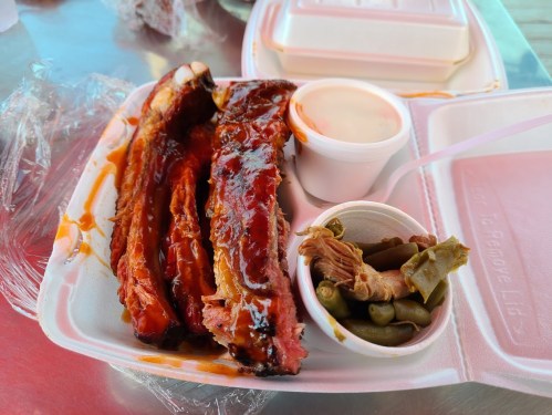 A takeout container with BBQ ribs, a small cup of sauce, and a side of pickled vegetables.