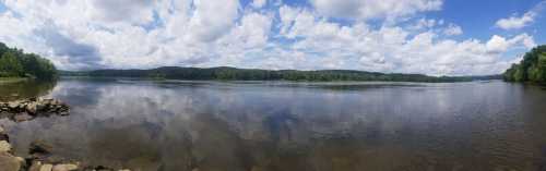 A serene river scene with lush green trees, blue skies, and fluffy clouds reflecting on the water's surface.