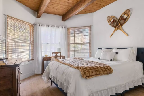 Cozy bedroom with a white bed, wooden beams, and snowshoes decor on the wall, featuring natural light from large windows.