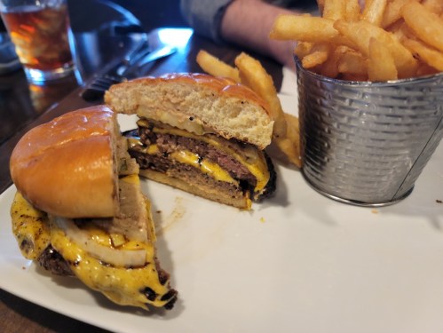 A close-up of a burger with layers of meat and cheese, served with a side of crispy fries in a metal cup.