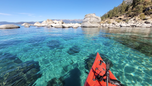 A red kayak floats on clear blue water, revealing rocky formations beneath the surface and a scenic shoreline in the background.