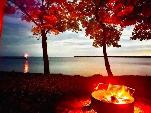A serene lakeside scene at dusk, featuring a glowing campfire and trees, with a distant lighthouse visible across the water.