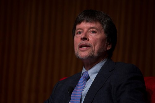 A man with a beard speaks into a microphone, wearing a suit and tie, against a wooden backdrop.