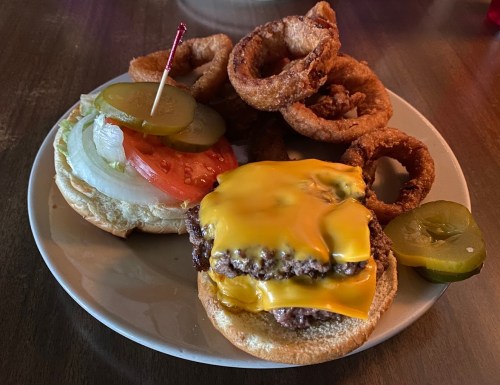 A plate with a cheeseburger topped with lettuce, tomato, and pickles, accompanied by onion rings and pickles.