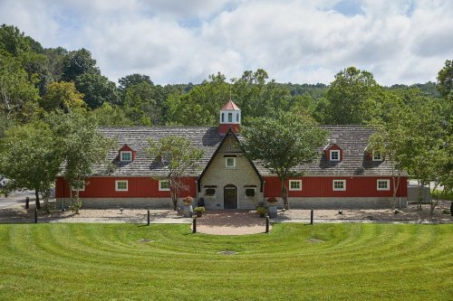 A red barn with a stone entrance, surrounded by trees and a manicured lawn under a cloudy sky.
