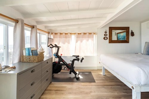 A bright, airy room featuring a bed, dresser, and exercise bike near large windows with light curtains.